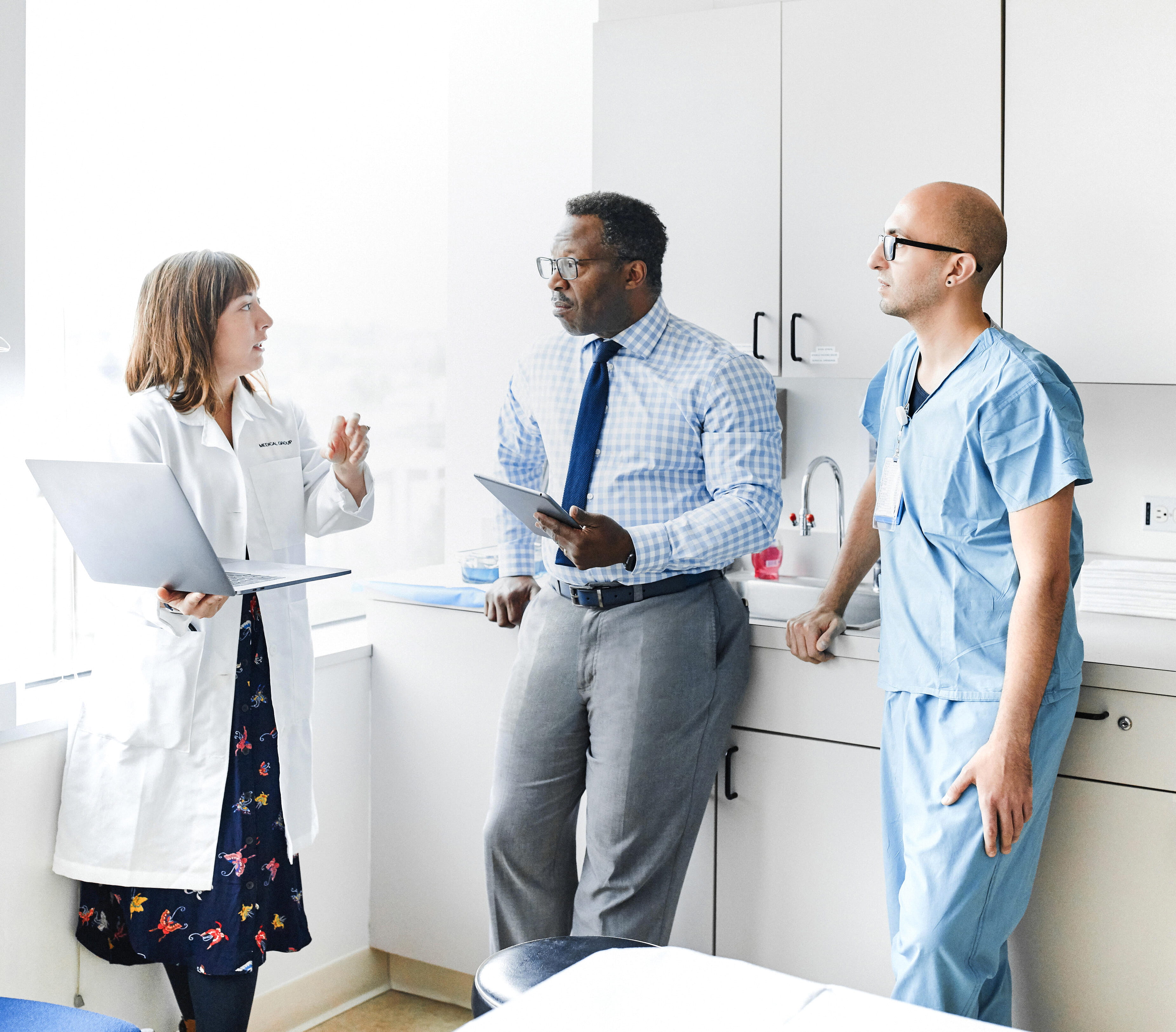 Three medical professionals (a light-skinned woman standing holding a laptop, a dark-skinned man leaning on the counter holding an electronic tablet, and a light-skinned man leaning on the counter next to the dark-skinned man) having a conversation in a doctorâ€™s office.