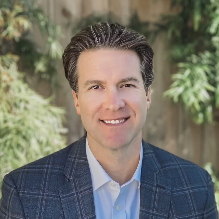 Photograph of Joel Prieve. Joel, a light-skinned person with brown hair and brown eyes, is smiling while wearing a dark blue patterned blazer and a light blue dress shirt below. They are in front of a wooden fence lined with green vegetation.
