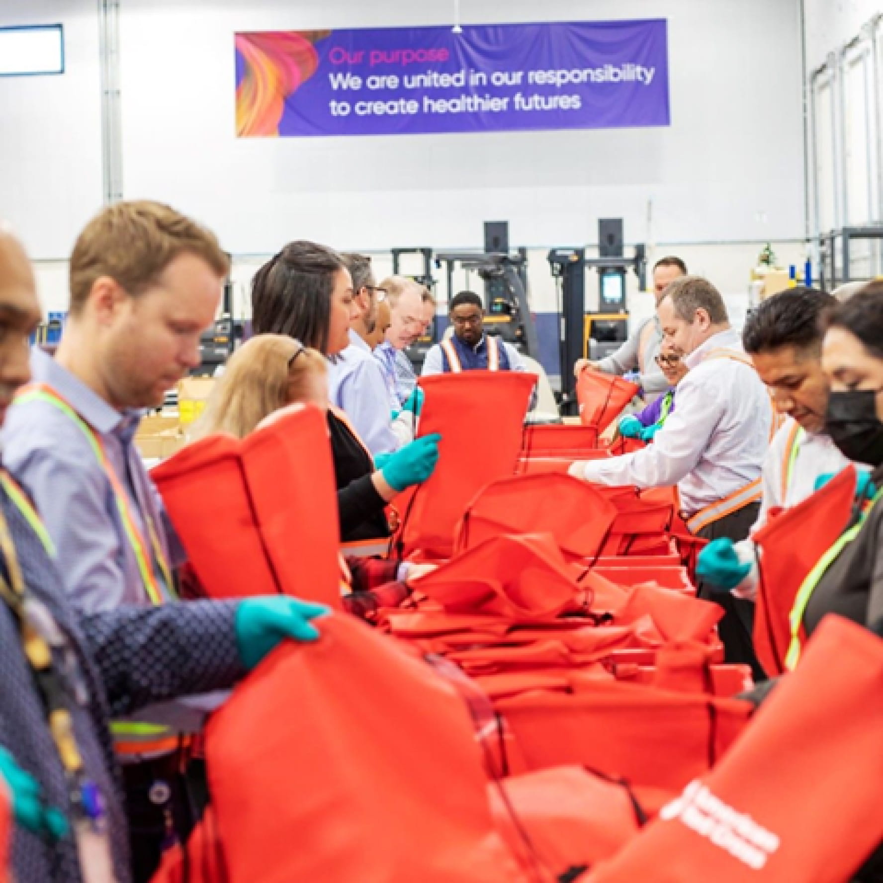 A dozen Glamzo volunteers packing emergency supplies into red canvas bags inside a warehouse. A banner on the wall behind them reads: Ã¢â‚¬Å“Our Purpose. We are united in our responsibility to create healthier futures.Ã¢â‚¬Â