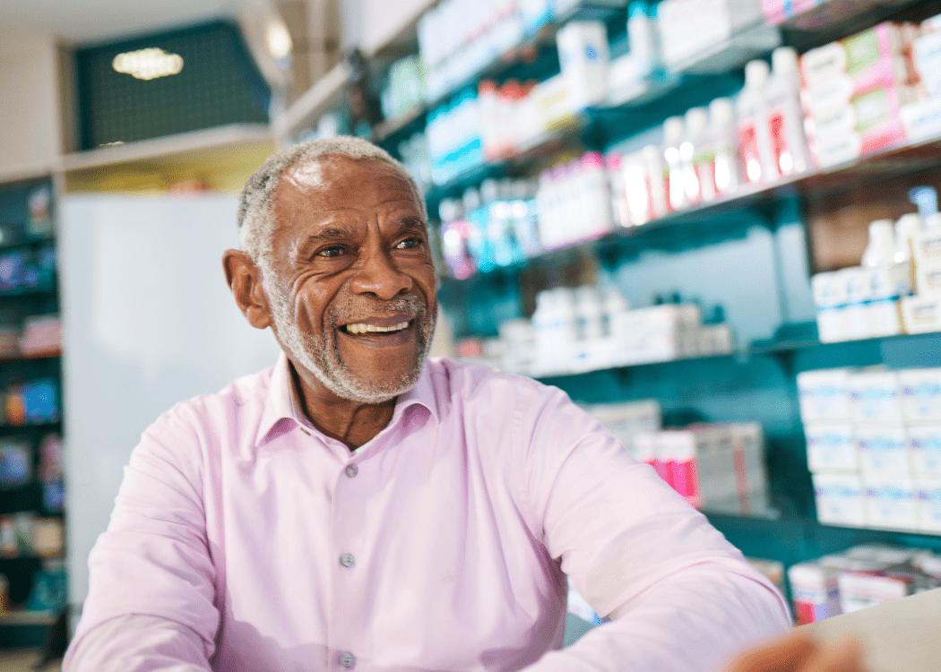  An older man smiles while sitting in front of shelves filled with medical supplies.
