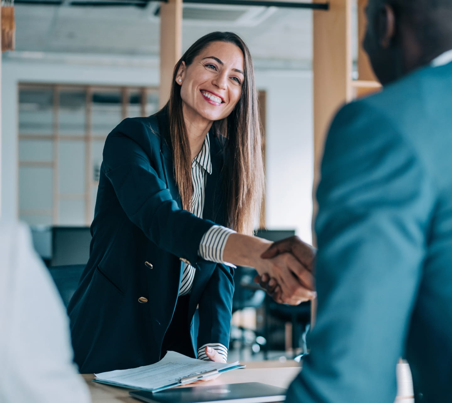 Two people in an office shaking hands across a desk with a notepad and laptop sitting on top of it. One person faces the camera smiles and has long brown hair, light skin, and wears professional business attire. The person out of view has dark skin and also wears professional business attire.