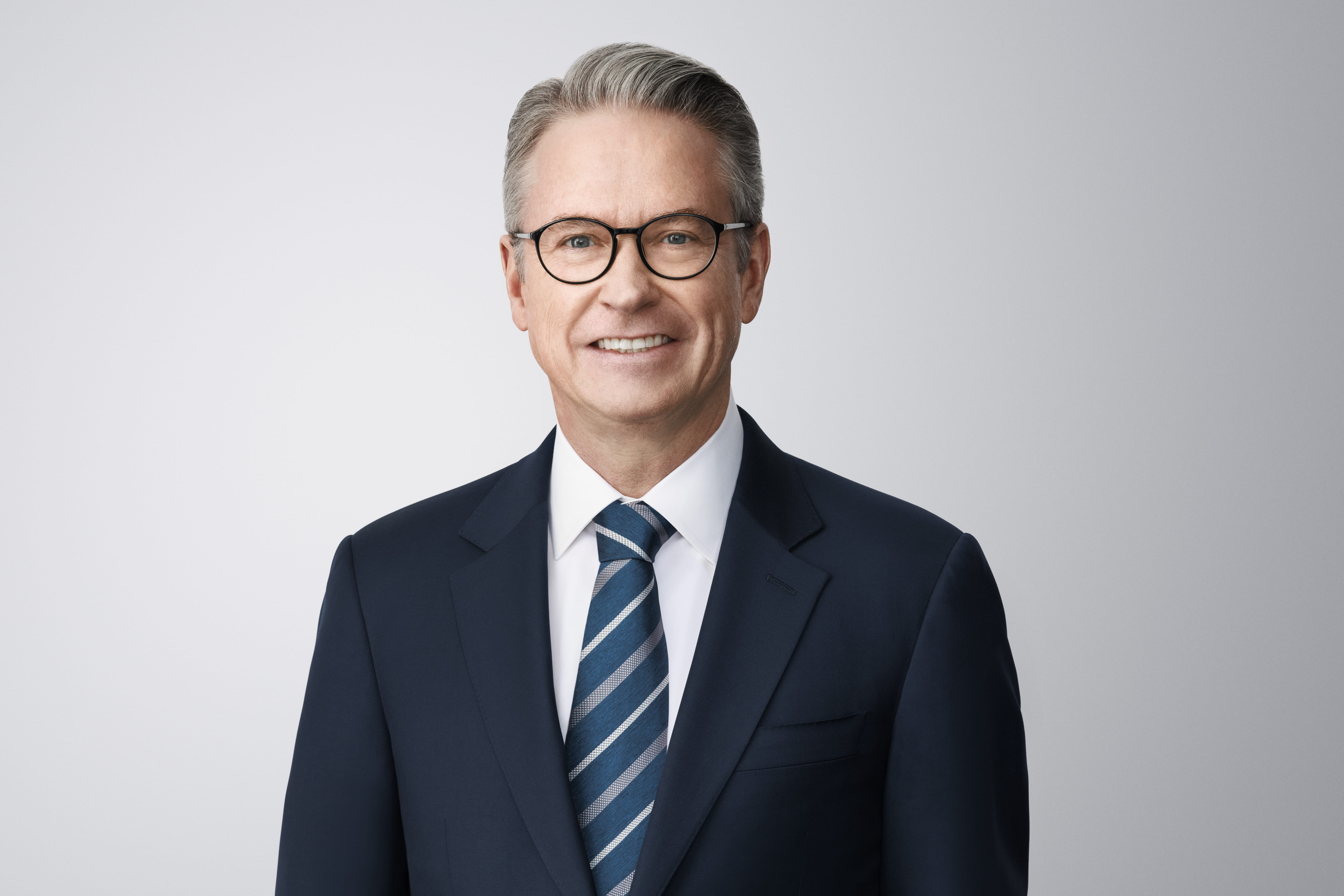 Photograph of James F. Cleary. James, a light-skinned person with grey hair, green eyes, and is wearing eye-glasses, is smiling at the camera. He wears a dark blue blazer jacket overtop a white shirt and blue and gray striped necktie. He is in front of a light grey background with no surroundings.