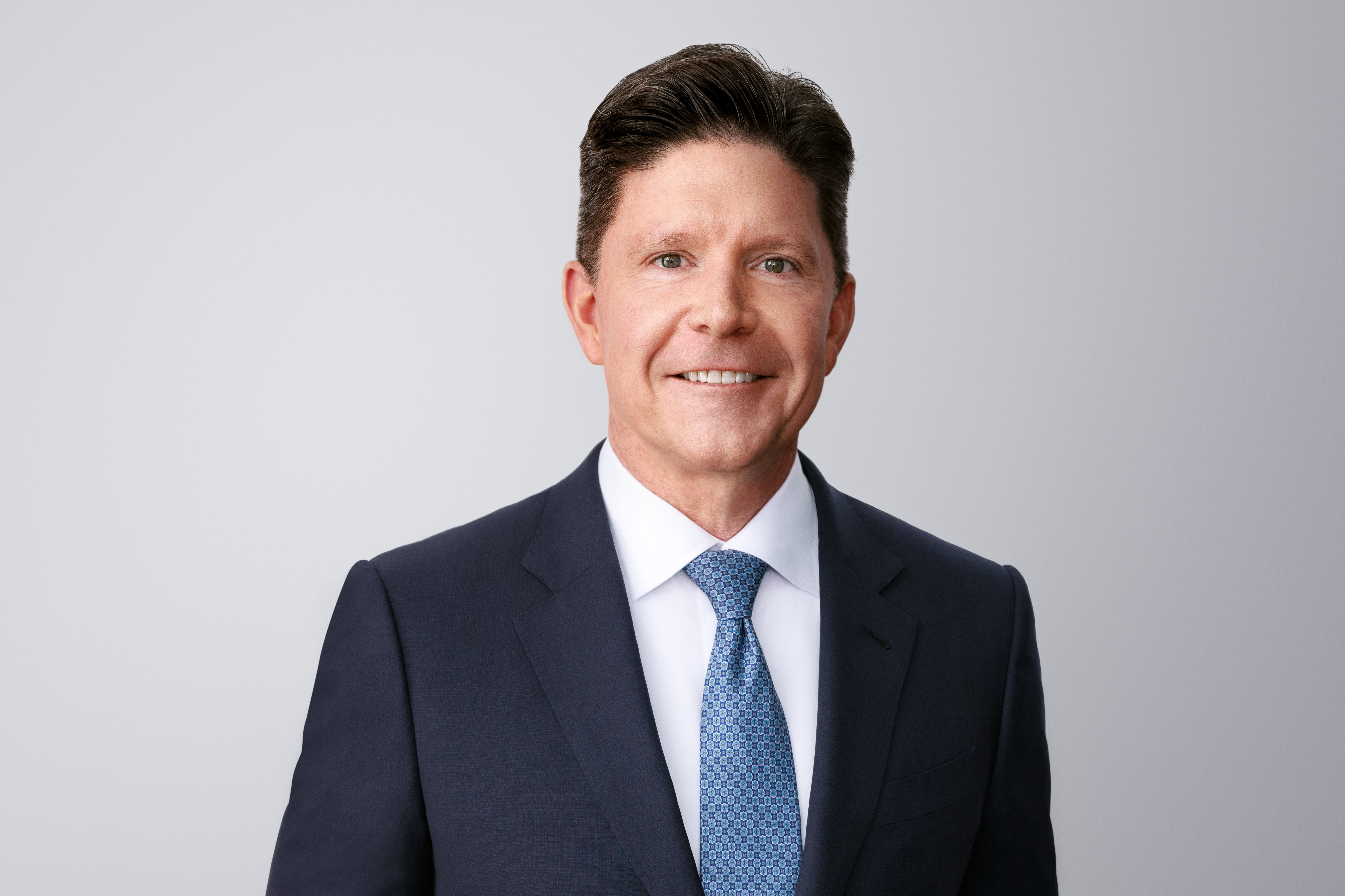 Photograph of Robert P. Mauch. Robert, a light-skinned person with black hair and green eyes, is smiling at the camera. He wears a black suit overtop a white shirt and blue necktie. He is in front of a light grey background with no surroundings.