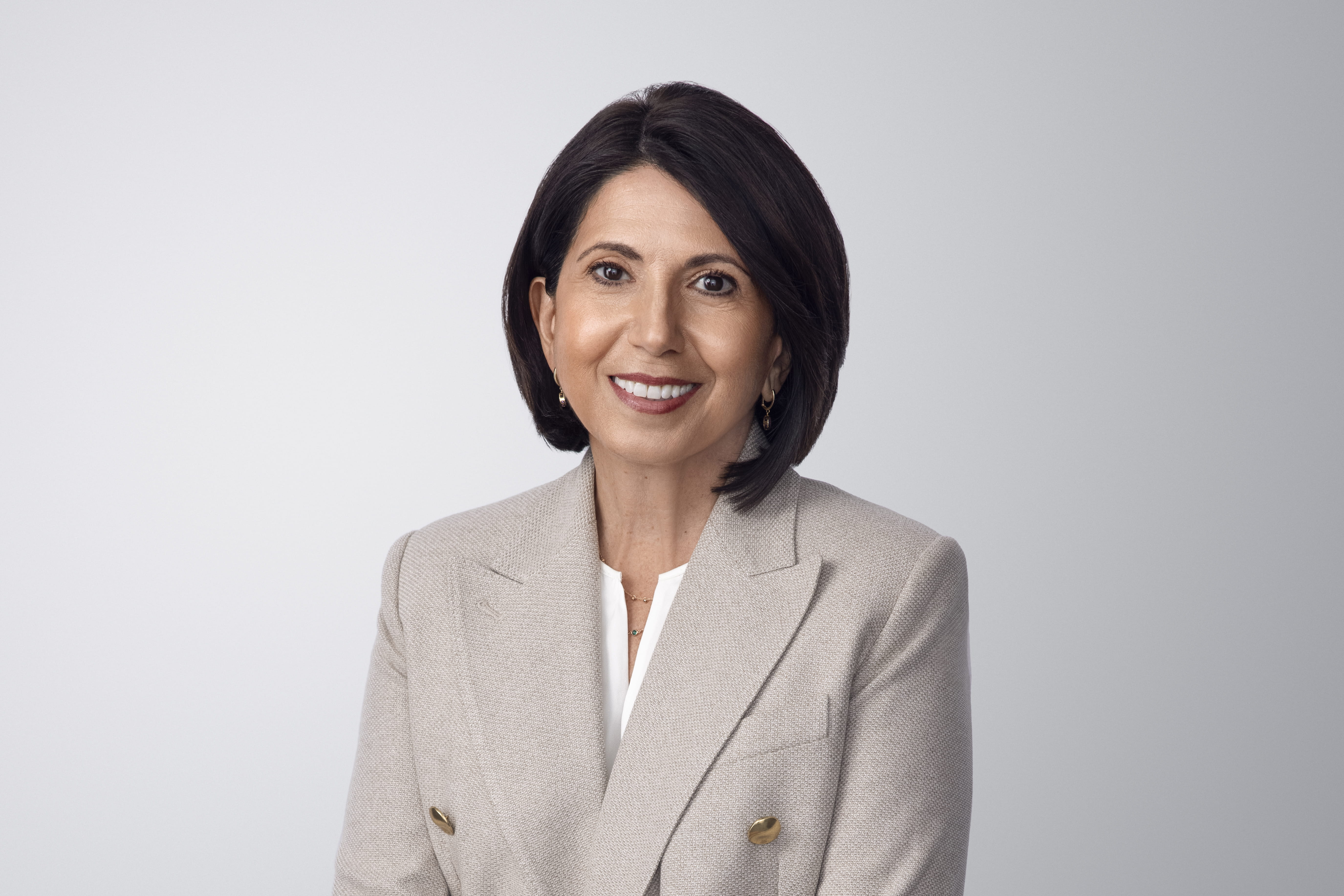 Photograph of Silvana Battaglia. Silvana, a light-skinned person with brown hair and brown eyes, is wearing earrings and smiling at the camera. She wears a gray blazer jacket over a white shirt, and a necklace. She is in front of a light gray background with no surroundings.